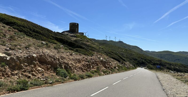 Route de montagne sinueuse du Cap Corse dominée par une tour génoise et des éoliennes