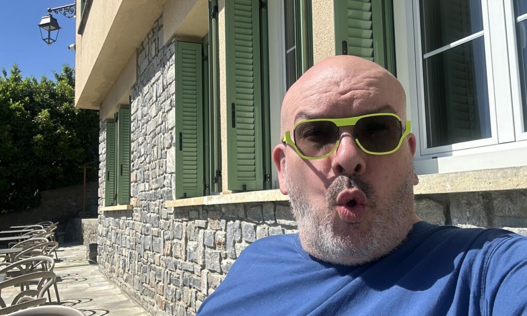 Selfie d'un homme chauve à lunettes vertes devant une maison aux volets verts