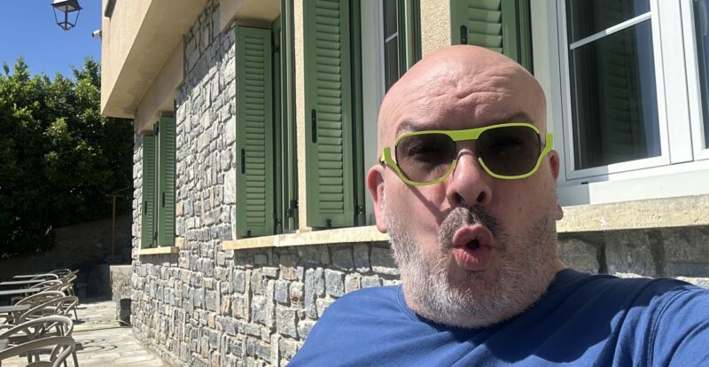 Selfie d'un homme chauve à lunettes vertes devant une maison aux volets verts