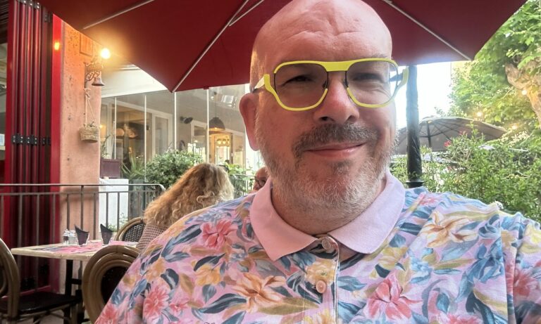 Homme souriant aux lunettes jaunes et chemise fleurie en terrasse sous parasol rouge