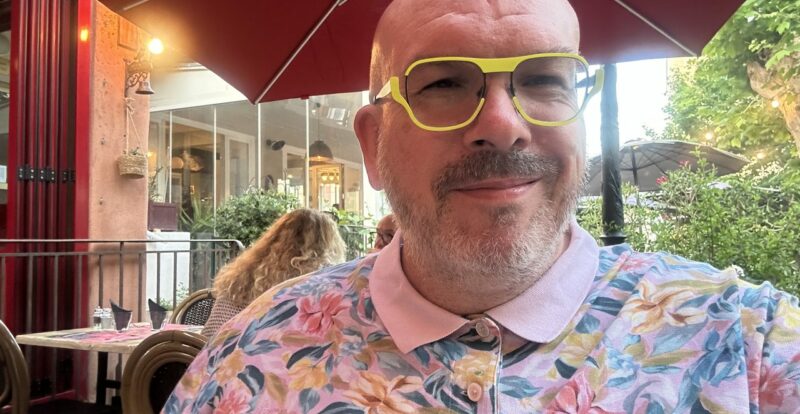 Homme souriant aux lunettes jaunes et chemise fleurie en terrasse sous parasol rouge