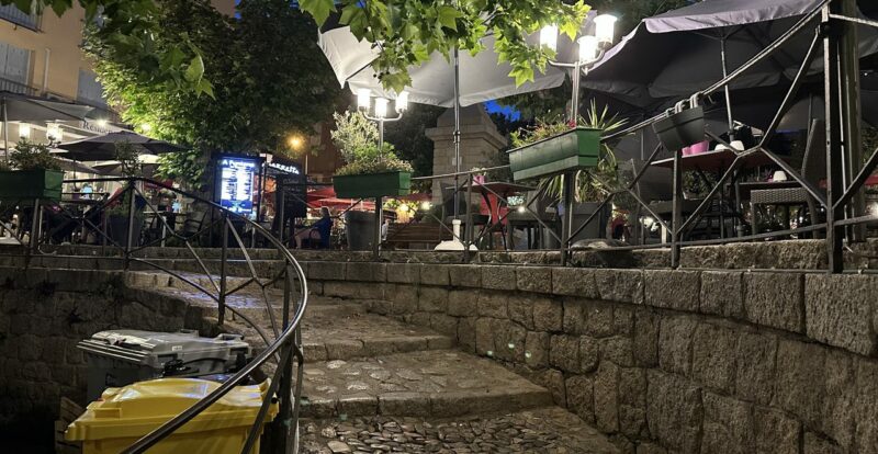 Ruelle pavée d'un village corse le soir avec terrasses éclairées et parasols