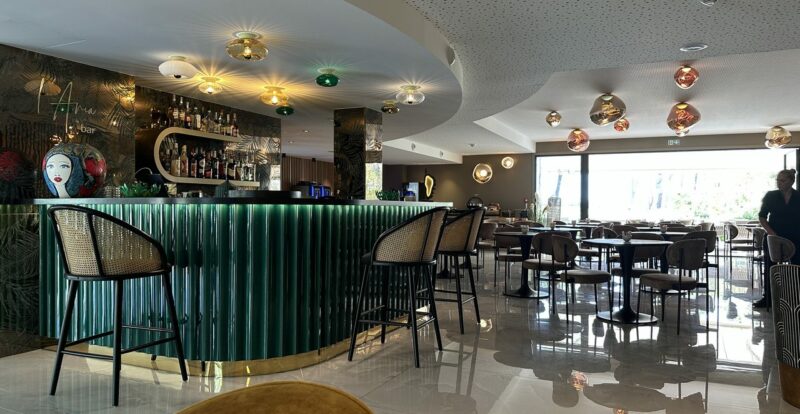 Bar d'hôtel design avec comptoir vert, suspensions cuivrées et chaises en rotin